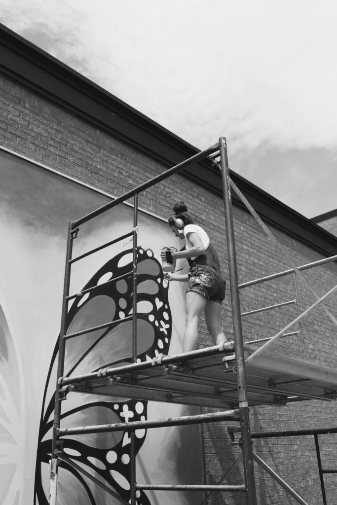 A black and white photo of a woman painting a butterfly on a wall.